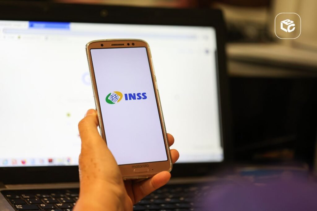 Mão segura celular com logo do INSS sobre notebook, em post sobre processo contra o INSS.