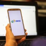 Mão segura celular com logo do INSS sobre notebook, em post sobre processo contra o INSS.
