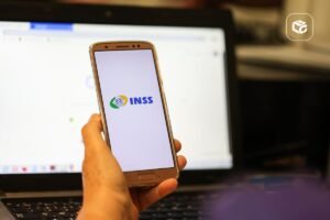Mão segura celular com logo do INSS sobre notebook, em post sobre processo contra o INSS.