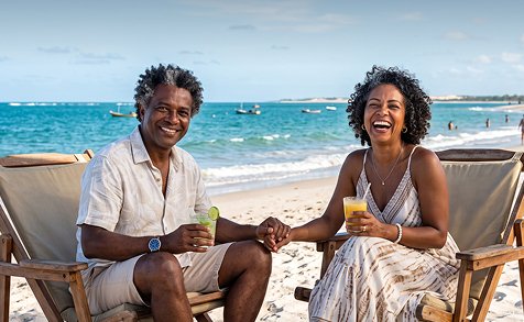 Casal sorrindo em cadeiras de praia, segurando bebidas, com o mar ao fundo.
